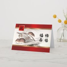 a card with two mice on it and chinese writing in the middle, sitting next to a vase filled with yellow flowers