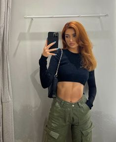 a woman taking a selfie in front of a mirror wearing green pants and a black crop top