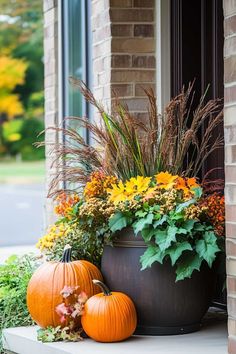 two pumpkins are sitting on the front porch next to some flowers and plants in a large planter