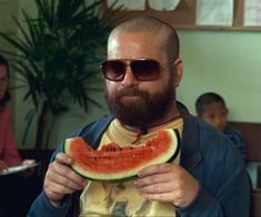 a man holding a slice of watermelon in front of his face with other people behind him