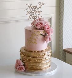 a pink and gold birthday cake with flowers on top is sitting on a white table