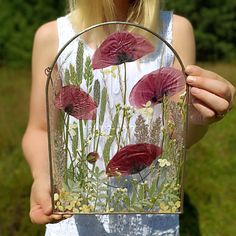 a woman is holding a glass vase with flowers in it and grass behind the bag