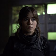 a woman standing in the dark with a black jacket and scarf on her shoulders, looking at the camera