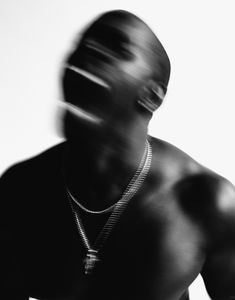 a man in black shirt and necklace standing next to a white wall with blurry image