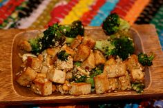 a wooden plate filled with tofu and broccoli on top of a colorful table cloth