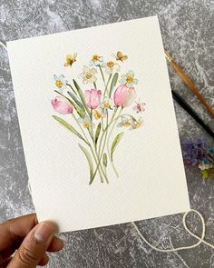 a person holding up a card with watercolor flowers on it next to some paintbrushes