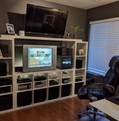 a room with a desk, chair, television and shelves filled with electronic equipment in it