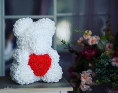 a white teddy bear with a red heart on it's chest sitting next to flowers