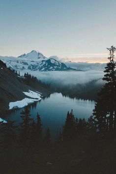 a lake surrounded by snow covered mountains and pine trees in the foreground with low clouds