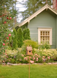 a birdhouse on a stick in front of a house with flowers and trees around it