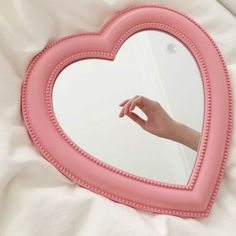 a pink heart shaped mirror hanging on a wall next to a white bed with a person's hand in it