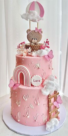 a pink cake decorated with clouds, flowers and a teddy bear