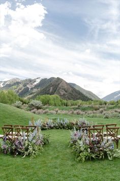 an outdoor ceremony set up with wooden chairs and flowers on the grass in front of mountains