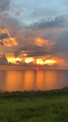 the sun is setting over the ocean with clouds in the sky and grass on the ground