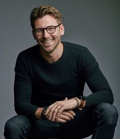 a man with glasses sitting on top of a wooden stool smiling at the camera while wearing a black sweater and jeans