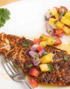 a white plate topped with fish covered in fruit and veggies next to a fork