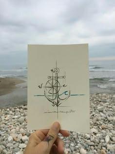 a hand holding up a piece of paper with an anchor on it in front of the ocean