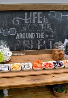 a wooden table topped with bowls filled with food next to a chalkboard sign that says life is better around the firepit