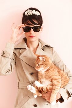 a woman in a trench coat holding a cat with sunglasses on it's head