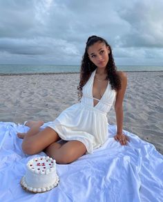 a woman sitting on top of a white blanket next to a cake in the sand