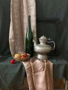 a table topped with a silver vase next to a basket of fruit and a bottle