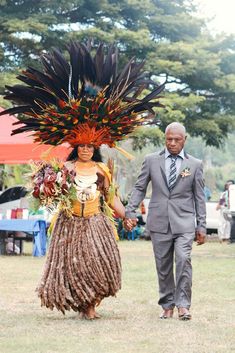 a man and woman dressed in native garb walking through the grass with flowers on their head