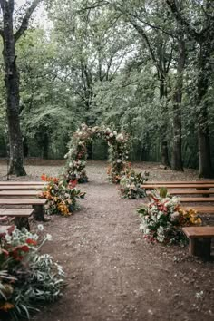 an outdoor ceremony setup with wooden benches and floral arrangements on the back wall, surrounded by trees