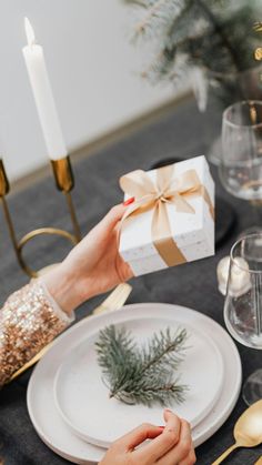 two hands holding a present over a white plate on a table with goldware and candles