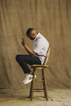 a man sitting on top of a wooden chair in front of a brown wall holding his hands together