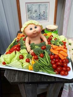 a table topped with a tray filled with lots of vegetables and a baby doll sitting on top of it