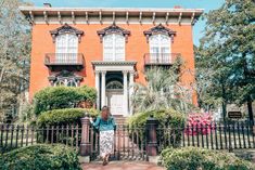 a woman standing in front of a red brick house with black iron fence and gate