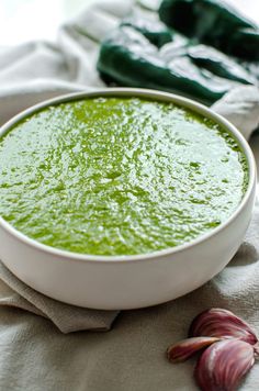 a white bowl filled with green soup next to garlic and an onion on a cloth