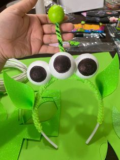 a hand holding up a green frog headband with googly eyes and an apple in the background