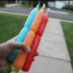 a person is holding four popsicles in front of the camera, and there are grass on the sidewalk behind them