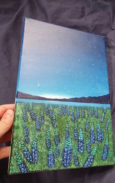 a person holding up a painting with blue flowers in the foreground and water on the far side