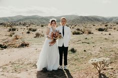 a bride and groom pose for a photo in the middle of an arid area with mountains behind them