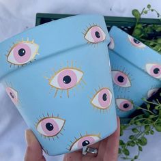 two blue pots with pink and yellow eyes painted on them are held up by someone's hand