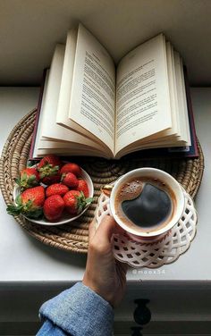 a person holding a cup of coffee next to an open book and bowl of strawberries