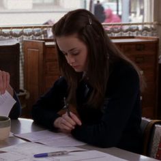 a woman sitting at a table with paper and scissors in her hand, writing on a piece of paper