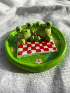 three little frogs sitting on top of a green plate with red and white checkered tablecloth