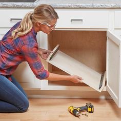 a woman kneeling down in front of a cabinet holding a piece of wood with a screwdriver next to it