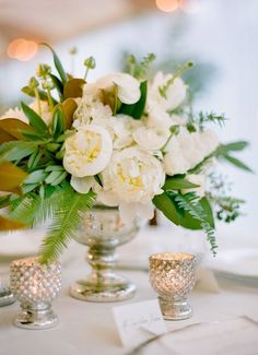 a vase with white flowers and greenery on top of a table next to candles