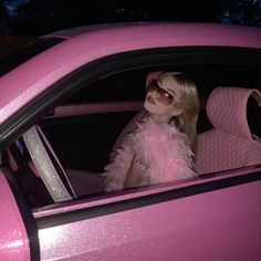 a woman sitting in the drivers seat of a pink car wearing sunglasses and a feather boa