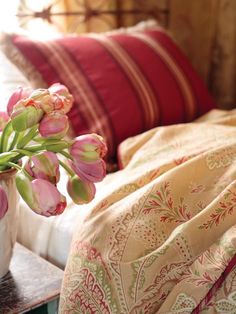 pink tulips in a vase on a table next to a bed with red and white pillows