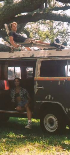 two people sitting on the roof of an old vw bus under a large tree