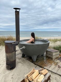 a woman is sitting in an outdoor bathtub on the beach with firewood nearby