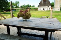 a wooden table sitting on top of a wooden deck next to a barn with a vase filled with flowers