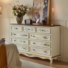 a white dresser with flowers on it in a bedroom next to a painting and lamp