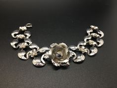 "This pretty bracelet is made of very lightweight silver-tone metal.  The center focal point is a raised rose with a rhinestone center.  Either side of the rose are silver leaves with small flowers with rhinestone centers.  It has a circular slide lock closure.  It measures 6 1/2\" long and about 1\" wide and is in beautiful condition as shown." Elegant Silver Flower Bracelets, Elegant Silver Bracelet With Rose Design, Elegant Silver Bracelets With Rose Design, Silver Flower Shaped Jewelry With Rose Design, Silver Floral Jewelry With Rose Design, Silver Flower Jewelry With Rose Design, Silver Rose Design Jewelry For Formal Occasions, Silver Flower Costume Jewelry, Silver Metal Jewelry With Rose Design