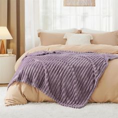 a bed with a purple blanket on top of it next to a lamp and window